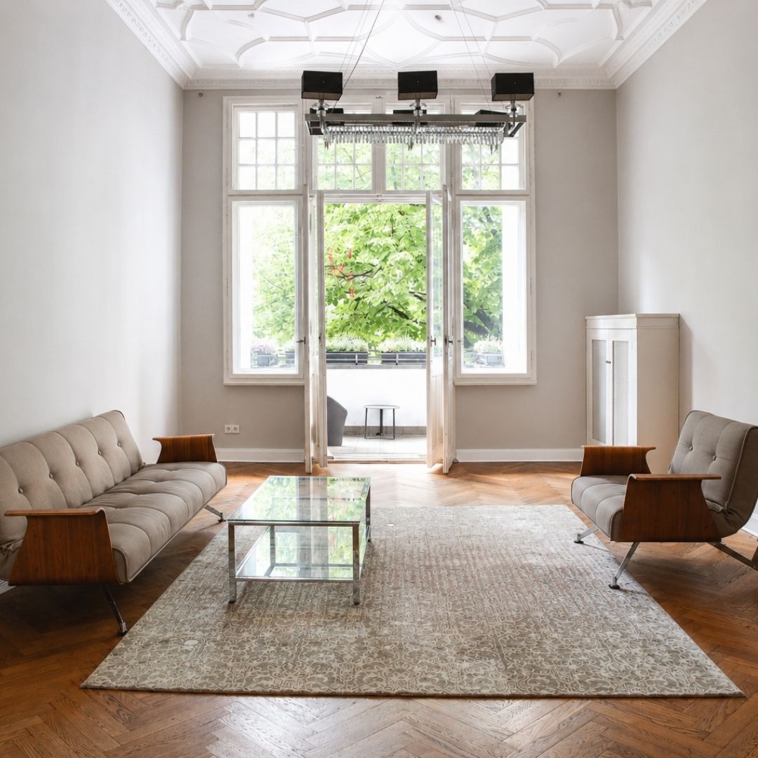 Lounge-Bereich von Beyond & Bloom Studio 51 mit hohen Altbaufenstern, Balkonblick, Stuckdecke, Parkettboden, Teppich, Sofa, Sessel und Glastisch.